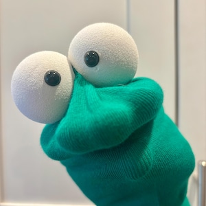 May include: A green sock puppet featuring two large, round, white foam eyes with small black pupils. The fabric of the puppet is a solid, vibrant green color with a soft texture, gathered around the base of the eyes to form a distinct shape.