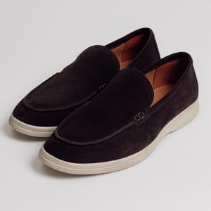 Op de afbeelding: Een paar donkerbruine su&egrave;de loafers met een ronde neus en een cr&egrave;mekleurige zool. De schoenen hebben een klassiek instapontwerp met gestikte rand en een leren binnenkant.