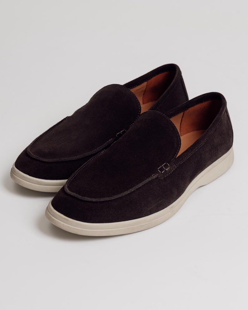 Op de afbeelding: Een paar donkerbruine su&egrave;de loafers met een ronde neus en een cr&egrave;mekleurige zool. De schoenen hebben een klassiek instapontwerp met gestikte rand en een leren binnenkant.