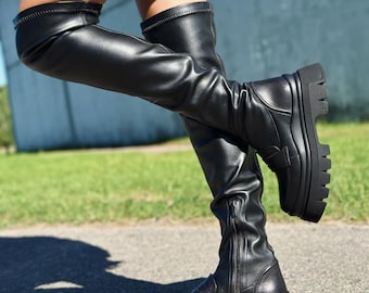 Op de afbeelding: Zwarte leren laarzen die boven de knie komen met een dikke platformzool en een zijrits. De laarzen hebben een strak, modern ontwerp en zijn perfect om een ​​stoere touch aan elke outfit toe te voegen.
