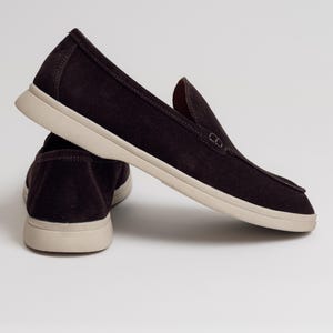 Op de afbeelding: Een paar donkerbruine instappers met een beige zool. De schoenen hebben een eenvoudig, klassiek ontwerp, met een ronde neus en een laag profiel. De bovenkant is gemaakt van een zacht, su&egrave;deachtig materiaal.