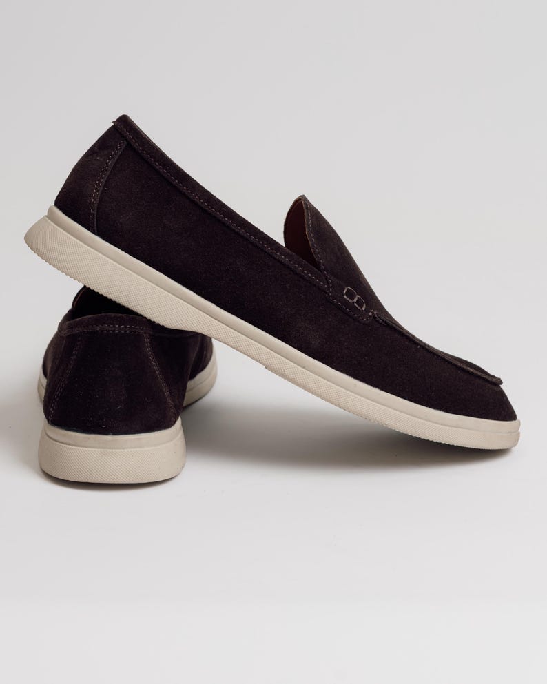 Op de afbeelding: Een paar donkerbruine instappers met een beige zool. De schoenen hebben een eenvoudig, klassiek ontwerp, met een ronde neus en een laag profiel. De bovenkant is gemaakt van een zacht, su&egrave;deachtig materiaal.