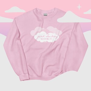 Może przedstawiać: Jasnoróżowa bluza z okrągłym dekoltem, ozdobiona centralną grafiką przedstawiającą białe chmury z napisem "pink clouding" wykonanym kursywą oraz mniejszym, bezszeryfowym tekstem "FROM THE THRILL OF IT" nadrukowanym na klatce piersiowej.