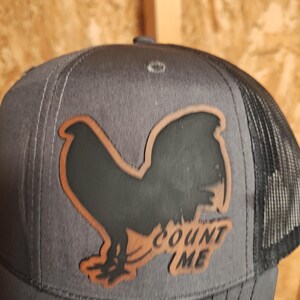 May include: A gray trucker hat with a black mesh back and a brown and black rooster design. The text "COUNT ME" is below the rooster. The hat has a curved brim and a snap closure.