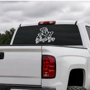 May include: A white vinyl decal applied to the rear window of a white pickup truck. The decal features a cartoon character wearing a chef hat and a chain, holding a money bag with a dollar sign, accompanied by the text Dough Boy in script font.