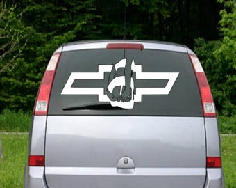 May include: A white car with a white decal on the back window. The decal is a silhouette of a person sitting on a bowtie. The bowtie is a common symbol for Chevrolet.