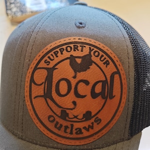 May include: A gray trucker hat with a black mesh back and a brown leather patch. The patch features the text "SUPPORT YOUR LOCAL OUTLAWS" in a circular design with a rooster graphic.