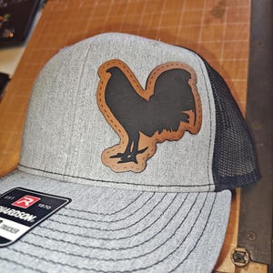 May include: Gray trucker hat with a black mesh back and a brown leather patch featuring a black rooster silhouette. The hat has a curved brim and a tag that reads "Richardson Trucker".