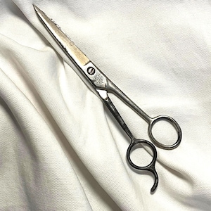 May include: A pair of silver and black barber shears with a curved handle and sharp blades. The shears are lying on a white textured surface. The blades have a serrated edge.