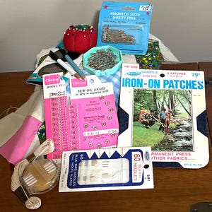 May include: A collection of sewing supplies including safety pins, needles, hooks, snaps, iron-on patches, and thread. The image features various packages and containers with sewing notions, perfect for crafting and mending projects.