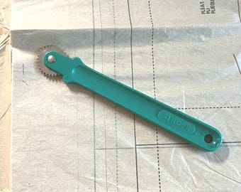 May include: A teal Singer tracing wheel with a serrated metal blade, resting on a sewing pattern. The handle is long and slender, with the Singer logo imprinted on it. The wheel is used for transferring pattern markings.