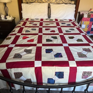 May include: A handmade quilt with a cream-colored background and a geometric pattern of burgundy and various patterned squares. The quilt is on a bed with white pillows and a dark metal frame. A smaller, colorful quilt is visible on a chair.
