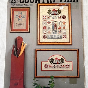 May include: A vintage "Country Fair" cross-stitch pattern book. The cover features framed cross-stitch samplers with country-themed motifs, including houses, hearts, and alphabets. A red wooden candle holder is on the left.