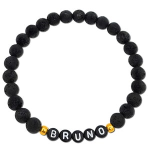Puede incluir: Pulsera de cuentas de roca volcánica negra con cuentas doradas y el nombre "BRUNO" escrito en letras blancas.