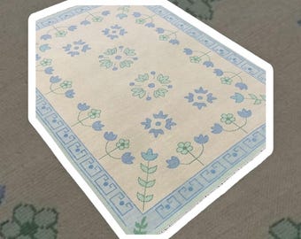 May include: A white rug with a blue and green floral pattern. The rug has a border of blue and white geometric shapes.