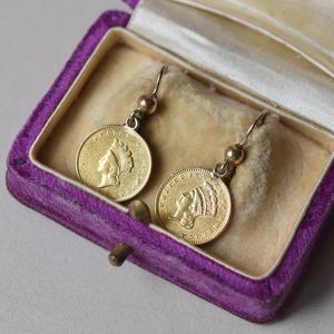 K&ouml;nnte beinhalten: Ein Paar goldene M&uuml;nzenohrringe mit dem Profil einer Frau auf jeder M&uuml;nze. Die Ohrringe befinden sich in einer Schmuckschatulle aus violetter Samt.