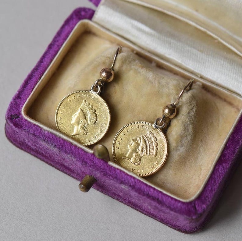 K&ouml;nnte beinhalten: Ein Paar goldene M&uuml;nzenohrringe mit dem Profil einer Frau auf jeder M&uuml;nze. Die Ohrringe befinden sich in einer Schmuckschatulle aus violetter Samt.