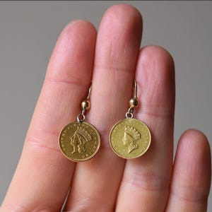K&ouml;nnte beinhalten: Ein Paar goldfarbene Ohrringe mit einem M&uuml;nzendesign, das das Profil einer Frau mit Krone zeigt. Die Ohrringe h&auml;ngen an einem silberfarbenen Haken.