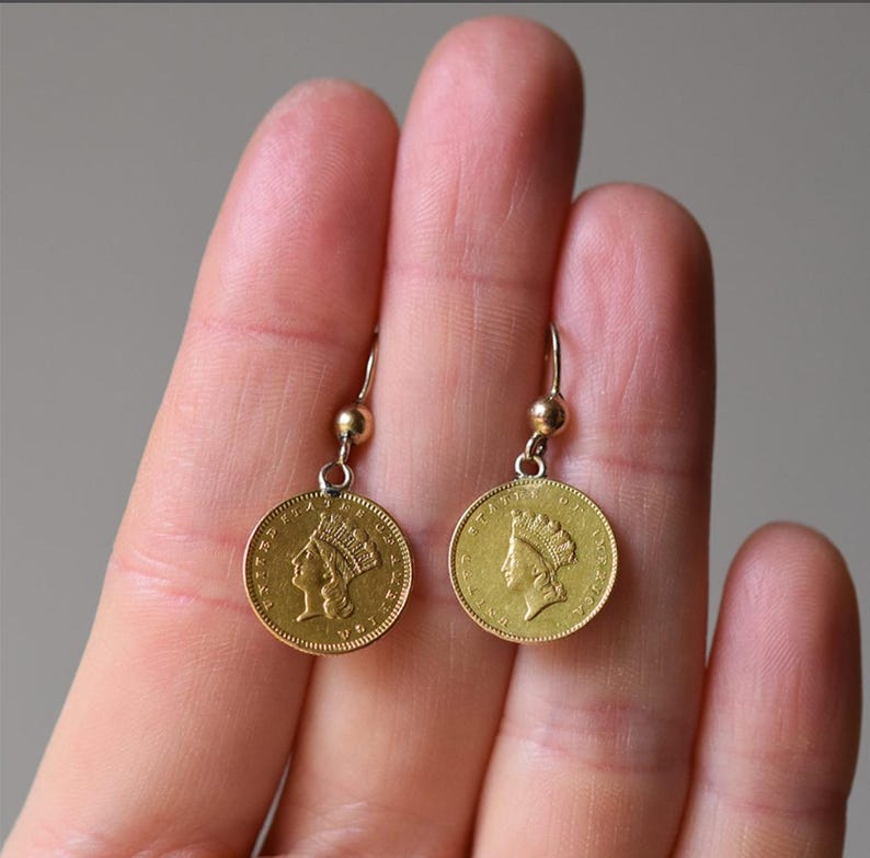 K&ouml;nnte beinhalten: Ein Paar goldfarbene Ohrringe mit einem M&uuml;nzendesign, das das Profil einer Frau mit Krone zeigt. Die Ohrringe h&auml;ngen an einem silberfarbenen Haken.