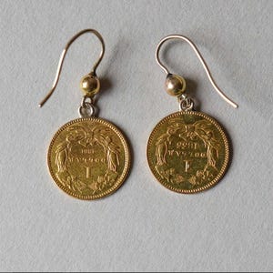 K&ouml;nnte beinhalten: Ein Paar goldene M&uuml;nzenohrringe mit einem silbernen Haken. Die M&uuml;nzen zeigen ein Design mit zwei V&ouml;geln mit einer Krone dar&uuml;ber und dem Jahr 1881. Die M&uuml;nzen sind mit einer kleinen goldenen Perle an den Haken befestigt.