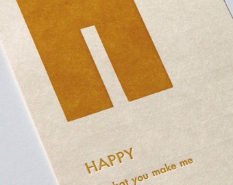 May include: A card with the word "HAPPY" in gold lettering on a cream background. The text "is what you make me" is also in gold lettering below the word "HAPPY".