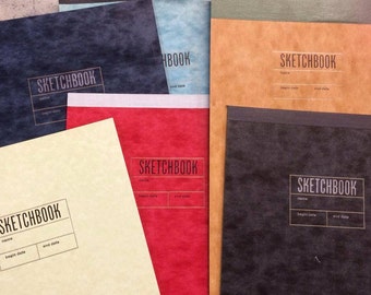 May include: A collection of sketchbooks in various colors, including navy, red, tan, and black. Each sketchbook features the word "SKETCHBOOK" in a rectangular box, with spaces for a name and dates.