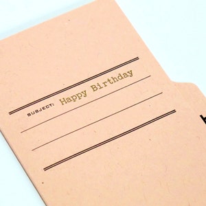 May include: A pink file folder with a gold inscription that reads "SUBJECT: Happy Birthday". The folder has a black tab with the letter "B" printed on it.