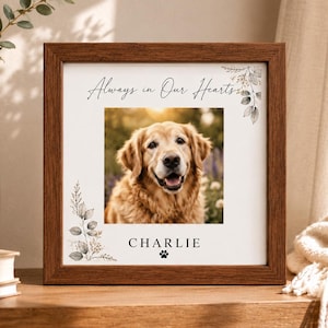 Op de afbeelding: Een vierkante houten lijst met een witte print met de tekst Always in Our Hearts en de naam CHARLIE onder een foto van een golden retriever. Het ontwerp bevat verfijnde botanische illustraties en een pootafdruk in de hoeken.