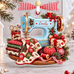 May include: A flat acrylic Christmas ornament shaped like a light blue sewing machine with the name Emily printed on the side. It features a stack of patterned fabric, a red pincushion, a gift box, and a red and white gingham ribbon bow.