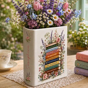 May include: A white ceramic book-shaped vase featuring a stack of classic book spines with titles like Little Women and Pride and Prejudice, surrounded by floral illustrations and filled with a bouquet of pink tulips and purple lavender.