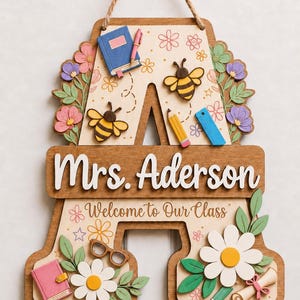 Könnte beinhalten: Ein Türschild aus Holz in Form des Buchstabens A mit dem Text Mrs. Aderson und Welcome to Our Class. Das Schild ist mit geschichteten Ausschnitten von Bienen, Blumen, einem Buch, einem Stift, einem Lineal, einer Brille und einer Schriftrolle verziert.