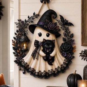 May include: A decorative wall hanging featuring a cream-colored ghost wearing a black witch hat and holding a black kitten. The ghost is surrounded by a wreath of black roses, leaves, and a small lantern, all suspended by a rope from a wall hook.