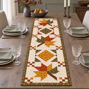 May include: A rectangular quilted table runner featuring a patchwork pattern of autumn maple leaves in shades of orange, yellow, brown, and green, bordered by a dark green floral print, centered on a wooden dining table with place settings.