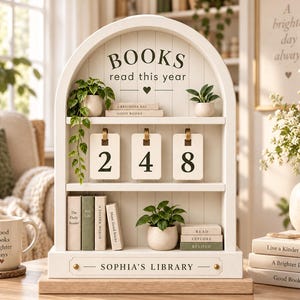 May include: A white arched wooden bookshelf display with three tiers. The top shelf features the text BOOKS read this year, a trailing green plant, and two small potted plants. The middle shelf displays three hanging number cards showing 248. The bottom shelf holds four small books and a potted plant. The base is labelled SOPHIA'S LIBRARY.