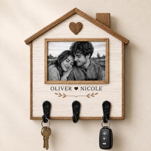 May include: A house-shaped wooden wall-mounted key holder with a white wood-grain finish and a dark wood frame. It features a central rectangular photo frame, a small heart cutout, and the text OLIVER HEART NICOLE with a leaf graphic. Three black metal hooks hold keys.