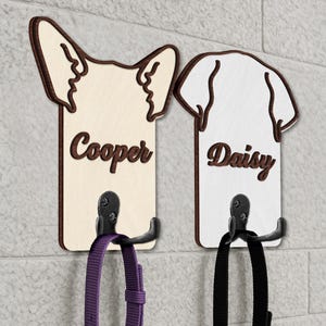 Op de afbeelding: Twee houten hondenriemhouders in de vorm van hondenkoppen, één bruin en één wit, met de namen "Cooper" en "Daisy" in bruin script. Elk heeft een zwarte haak met een halsband, één paars en één zwart. De houders zijn gemonteerd op een witte bakstenen muur.