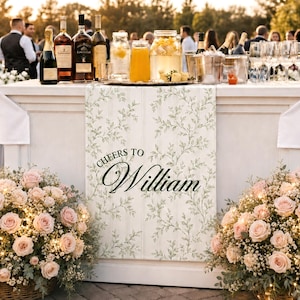 May include: A white bar table runner featuring a green botanical leaf pattern and the text Cheers to William in black script. The runner is draped over a white table, flanked by two large wicker baskets filled with pink roses and fairy lights.
