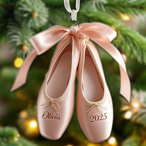 Könnte beinhalten: Ein Paar rosa Ballettschuhe mit einer großen passenden Schleife und einem weißen Band zum Aufhängen. Die Schuhe tragen den Namen "Olivia" und die Zahl "2025" in Gold gestickt. Der Hintergrund ist ein verschwommenes Bild eines Weihnachtsbaums.