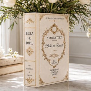 May include: A rectangular cream-colored ceramic vase shaped like a book, featuring gold ornate borders and text reading "A LOVE STORY," "Bella & David," "EST 16.08.2026," and "EVERY PAGE LEADS US HOME." It is filled with white tulips and greenery.