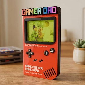 May include: A red picture frame shaped like a retro game console, with the text "GAMER DAD" in colorful pixelated letters. The frame holds a photo and includes the text "Coolest Dad in the world" and "GAME MASTER, HOME HERO".