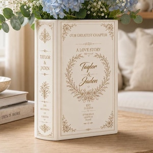May include: A rectangular cream-colored ceramic vase shaped like a book, featuring gold-toned decorative filigree and text that reads "Our Greatest Chapter," "A Love Story," "Taylor & Jaden," and "Est. 22.12.2026," filled with blue hydrangea flowers.
