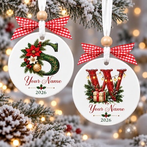 Könnte beinhalten: Zwei runde weiße Keramikornamente, die an weißen Bändern mit Holzperlen und rot-weiß karierten Schleifen hängen. Eines zeigt einen grünen Buchstaben S mit Weihnachtssternen, das andere einen roten Buchstaben W mit Lichterketten und einem Geschenk.