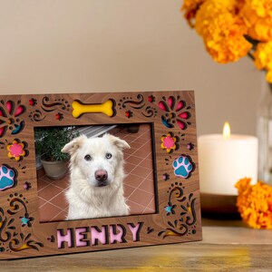 May include: A wooden photo frame with a picture of a white dog with blue eyes. The frame is decorated with colorful floral and paw print designs, and the dog's name, "HENRY," is displayed in pink letters.