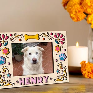 May include: A wooden picture frame with a photo of a dog named Henry. The frame is decorated with colorful floral and paw print cutouts. A yellow bone shape is at the top. The name "HENRY" is in pink letters at the bottom.