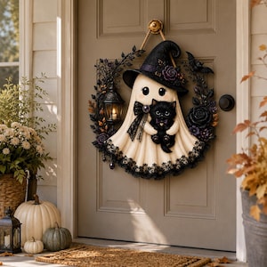 May include: A decorative wreath featuring a cream-colored ghost wearing a black witch hat and holding a small black kitten. The ghost is surrounded by black floral accents, a miniature lantern, and lace trim, all suspended by a rope on a door.