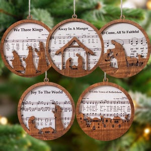 May include: Five wooden Christmas tree decorations. Each circular ornament has a cutout design of a nativity scene, with the text "We Three Kings", "Away In A Manger", "O Come, All Ye Faithful", "Joy To The World", and "O Little Town of Bethlehem" visible.