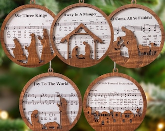 May include: Set of five wooden Christmas ornaments. Each ornament features a circular frame with a cutout design depicting a nativity scene. The text "We Three Kings", "Away In A Manger", "O Come, All Ye Faithful", "Joy To The World", and "O Little Town of Bethlehem" are visible.