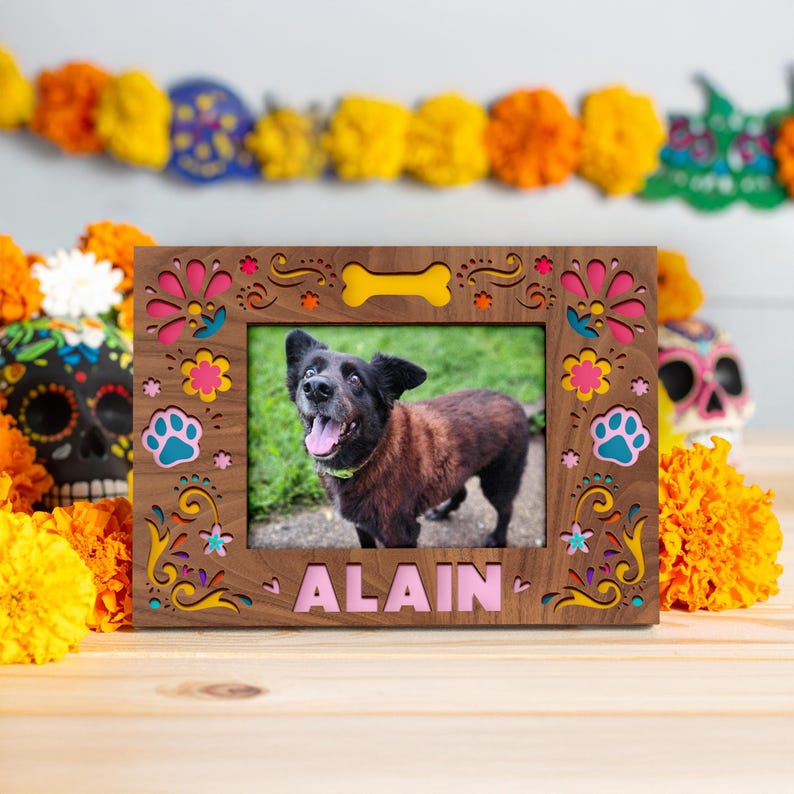 May include: Wooden photo frame with a picture of a dog named Alain. The frame is brown with colorful floral and paw print cutouts. A yellow bone shape is at the top. The background includes marigolds and a Day of the Dead skull.