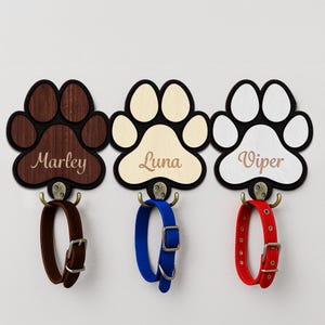 May include: Three paw-shaped wall hooks with pet names: "Marley", "Luna", and "Viper". Each paw has a different color scheme: brown, beige, and white. Each hook holds a dog collar in brown, blue, and red. The hooks are mounted on a white wall.