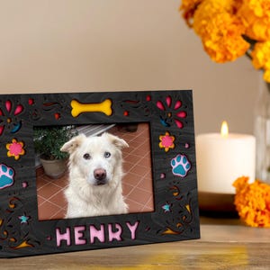 May include: A black wooden picture frame with colorful floral and paw print cutouts. The frame holds a photo of a white dog with the name "HENRY" in pink letters. A yellow bone cutout is at the top.